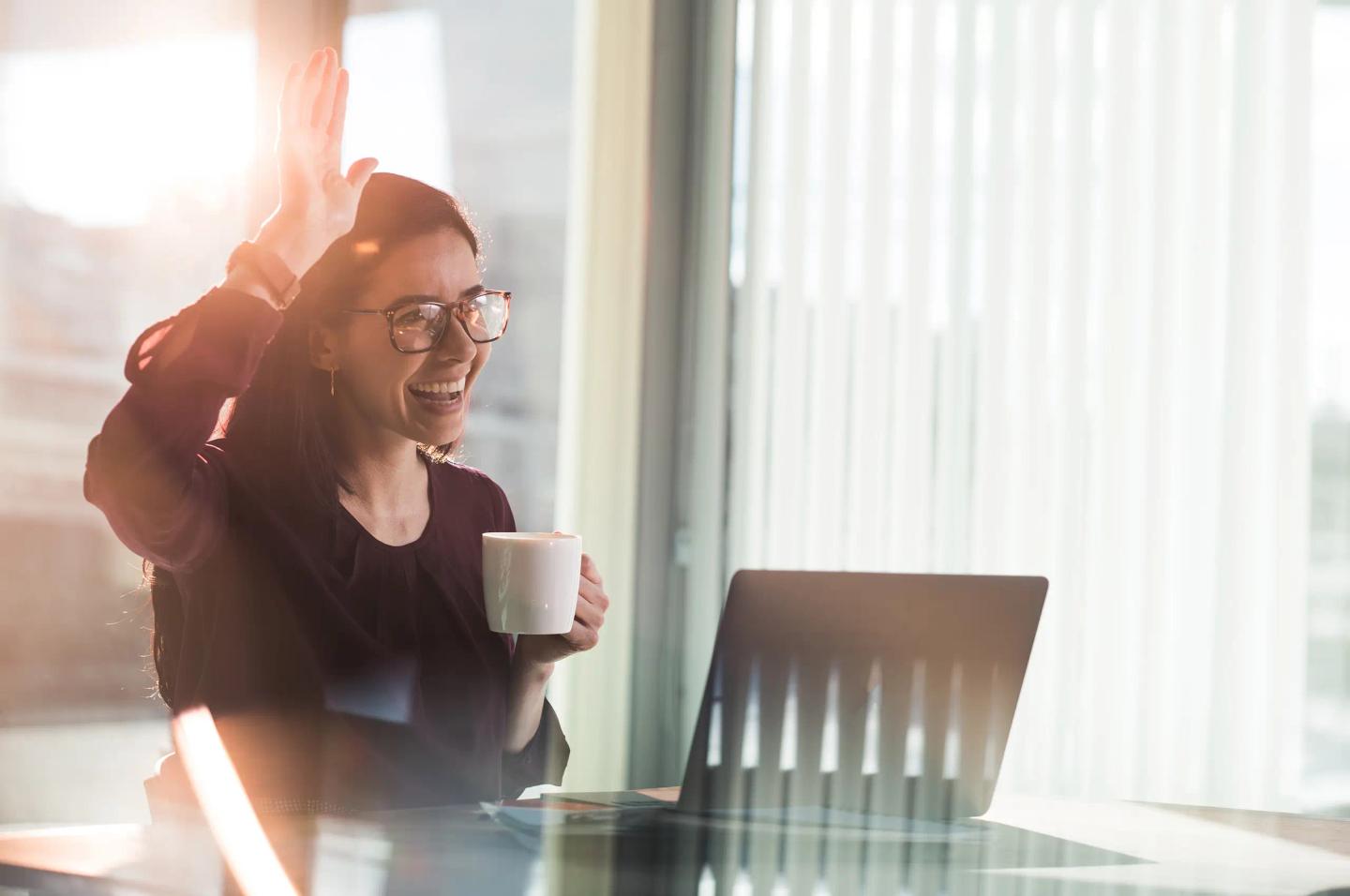 Leende person på jobbet med laptop och kaffekopp räcker upp en hand i hälsning till en kollega.