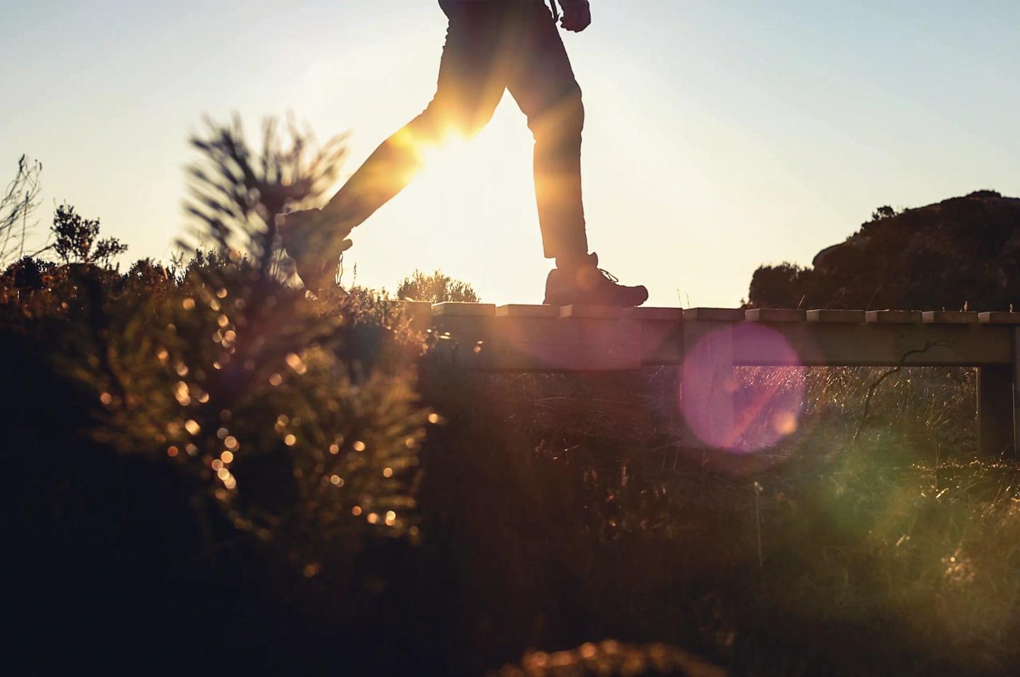 En person i vardagskläder går en promenad i det öppna landskapet och korsar en bäck över en enkel träbro.