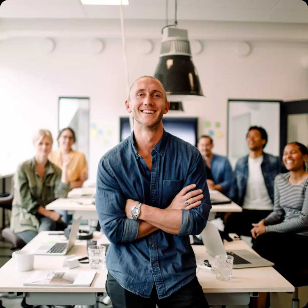 En nöjd chef ler mot kameran i kontorsmiljö med hela sitt team bakom sig.
