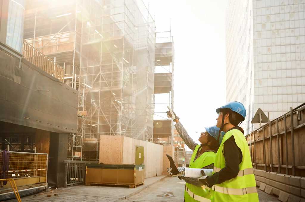 Två personer med hjälmar och reflexvästar inspekterar en ny byggnad under uppförande.